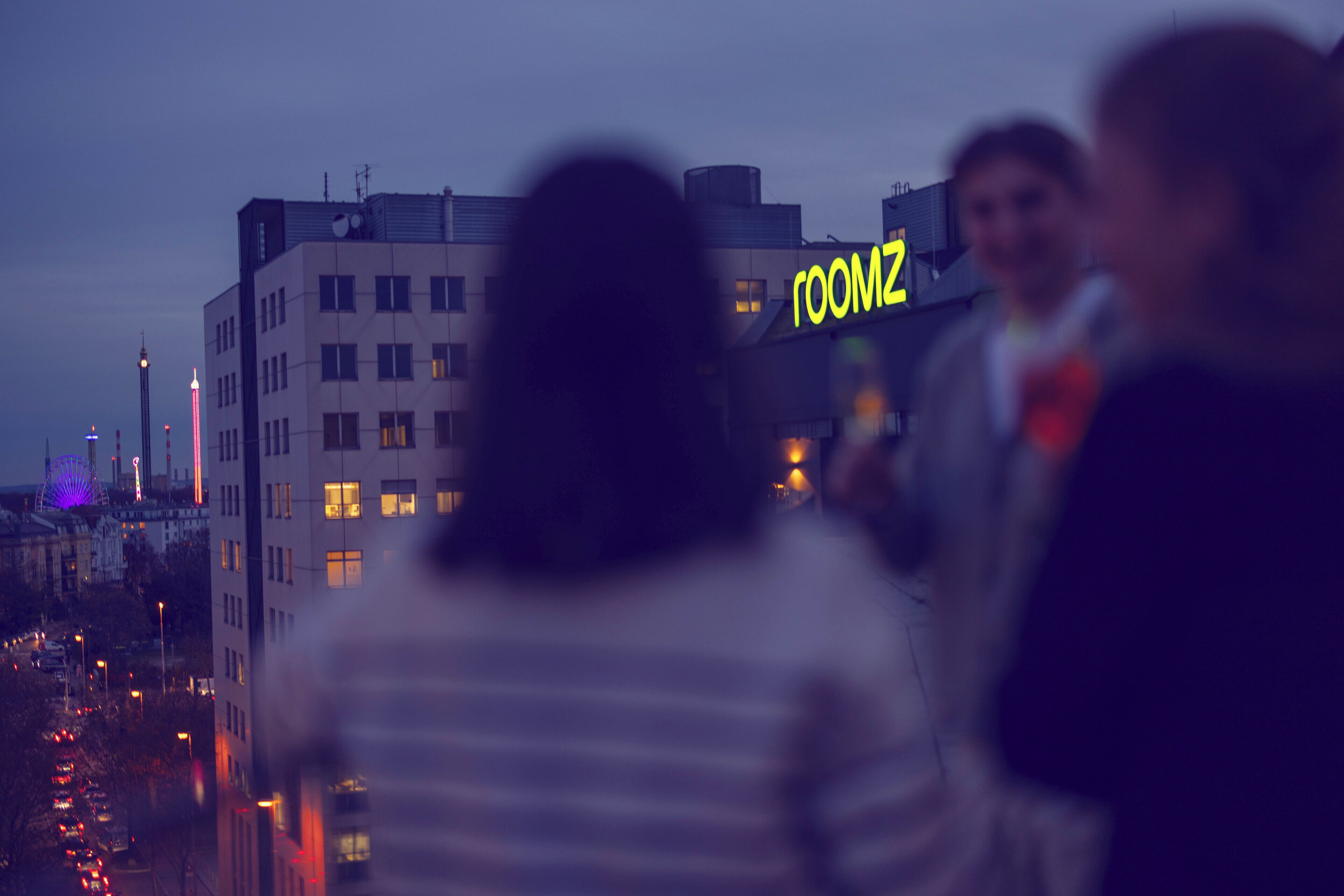 A building with a neon sign reading "ROOMZ" and a view of a cityscape with buildings and lights.