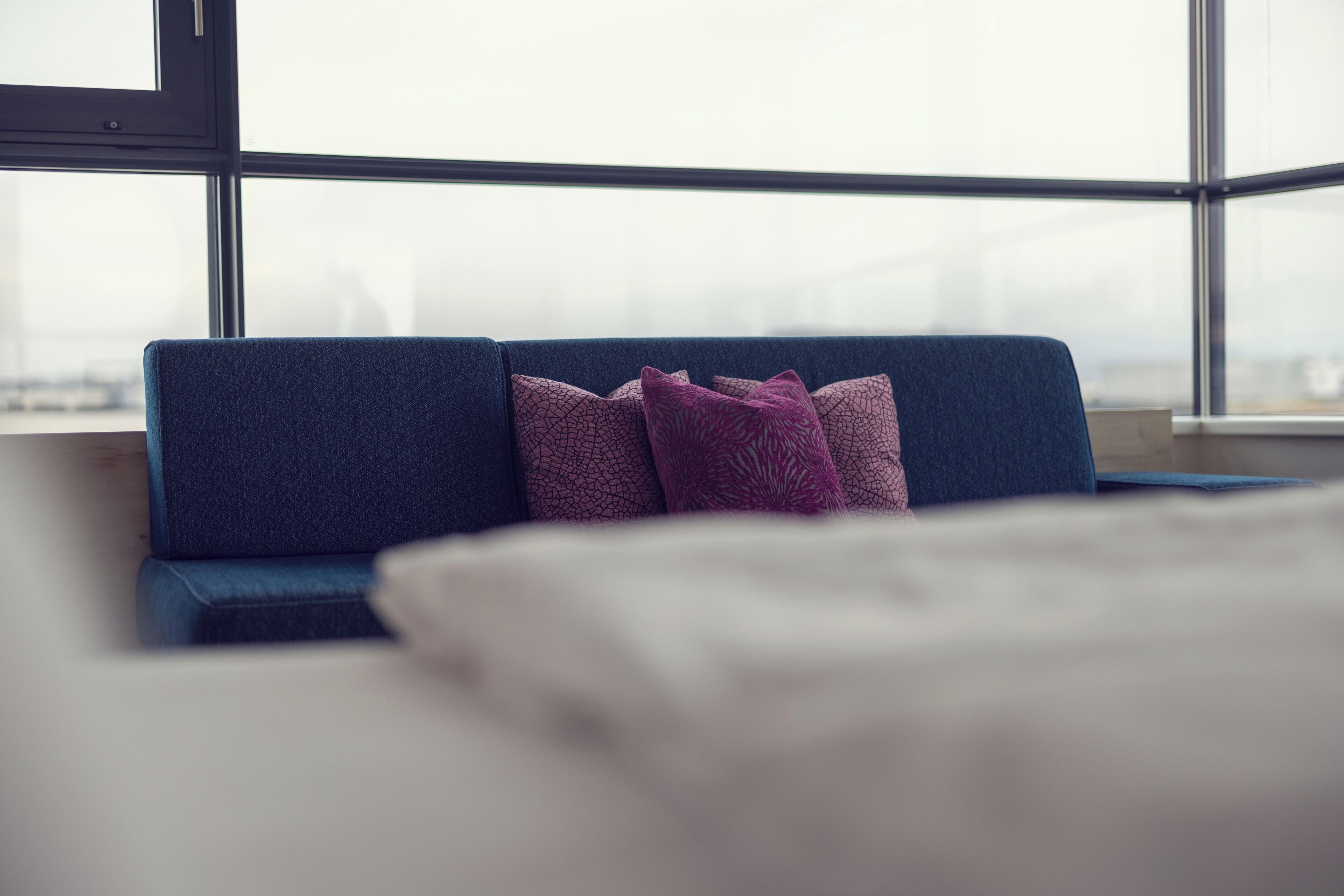 Quarto moderno com sofá azul, duas almofadas estampadas e uma janela ampla com vista para a paisagem urbana.