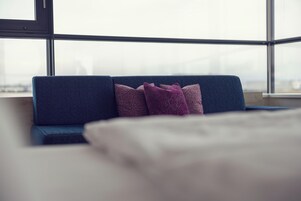 Quarto moderno com sofá azul, duas almofadas estampadas e uma janela ampla com vista para a paisagem urbana.