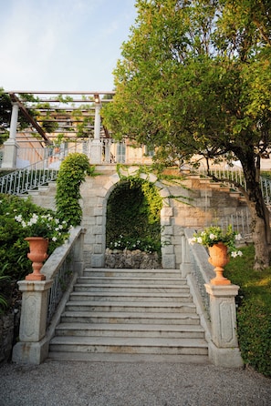A stone staircase leading up to a covered area with a metal railing, surrounded by greenery and a clear sky. - Hotel Villa Cipressi - by R Collection Hotels (Varenna)