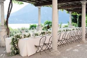An outdoor dining area with tables set for a meal, surrounded by columns and a view of a body of water and mountains. - Hotel Villa Cipressi - by R Collection Hotels (Varenna)