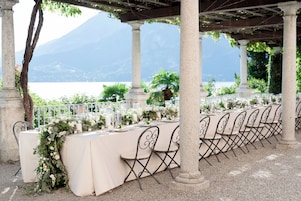 An outdoor dining area with tables set for a meal, surrounded by columns and a view of a body of water and mountains.