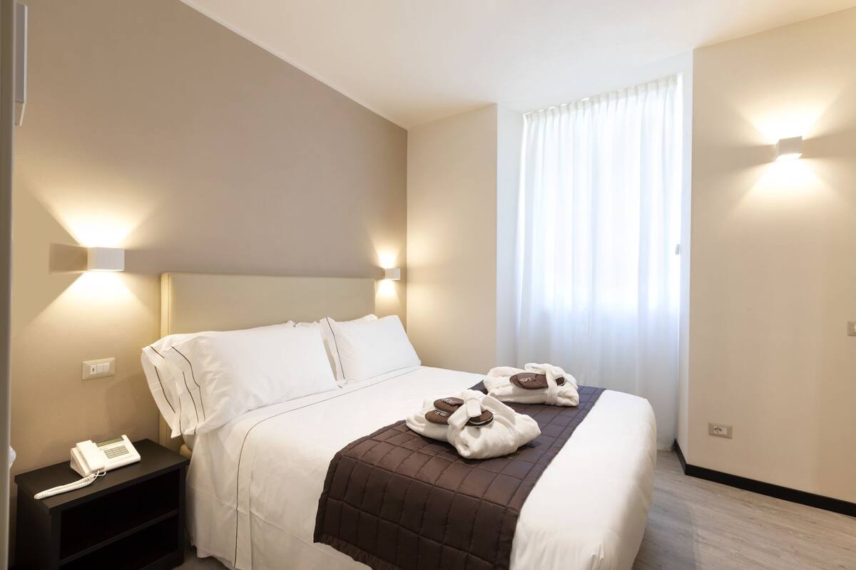 A hotel room with a bed, bedside table with a phone, and a window with curtains.