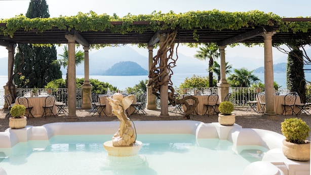 Zona de piscina con una fuente, rodeada de sillas y mesas, y vistas al lago y las montañas.