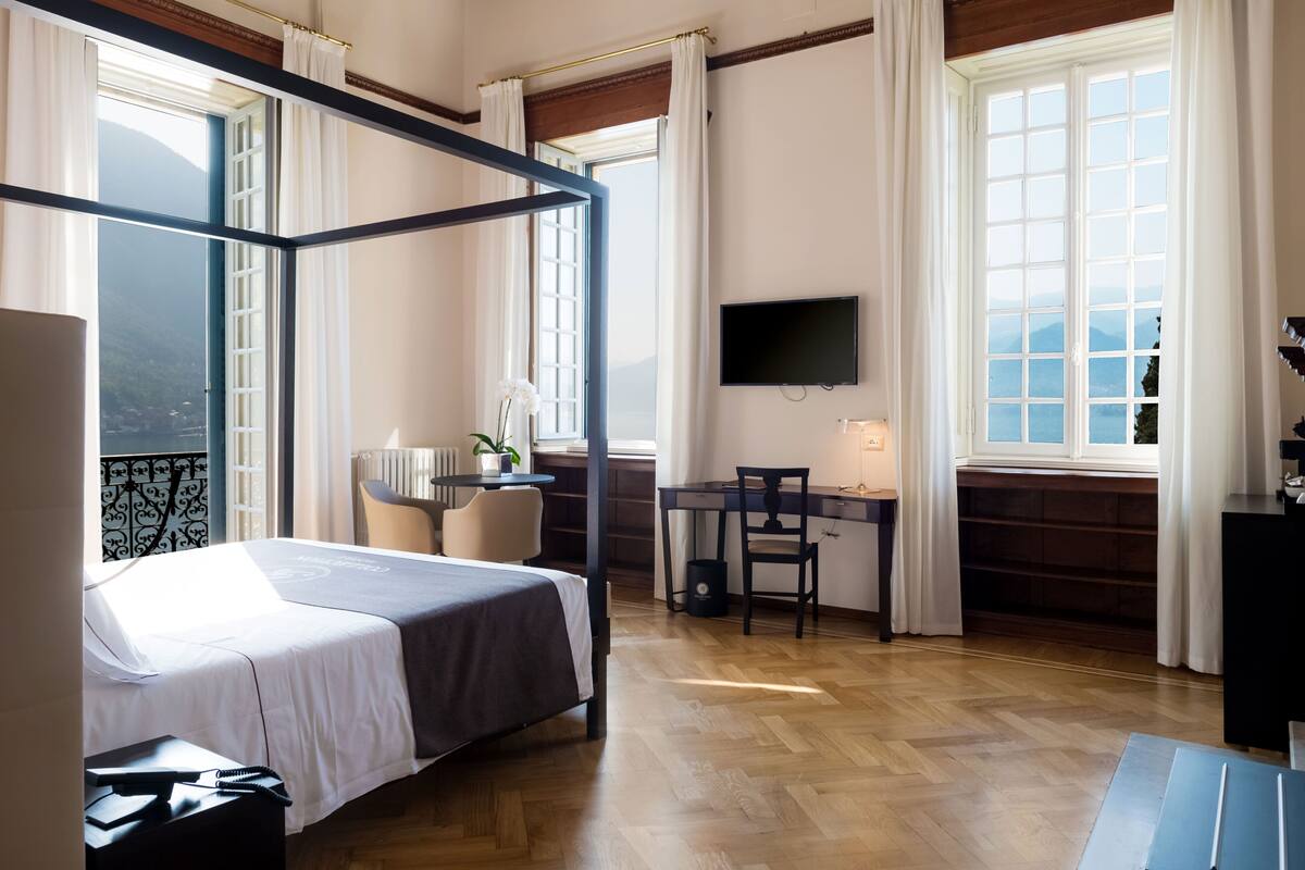 A room with a four-poster bed, a desk, a chair, a television, and large windows with curtains.