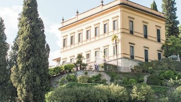 Ein großes, zweistöckiges Gebäude mit einer symmetrischen Fassade, gelegen an einem Hang mit einer Steinmauer und einer Brücke im Vordergrund.
