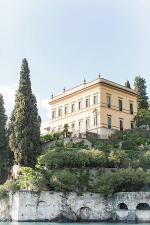 A large, two-story building with a symmetrical facade, situated on a hillside with a stone wall and a bridge in the foreground. - Hotel Villa Cipressi - by R Collection Hotels (Varenna)