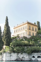 A large, two-story building with a symmetrical facade, situated on a hillside with a stone wall and a bridge in the foreground.