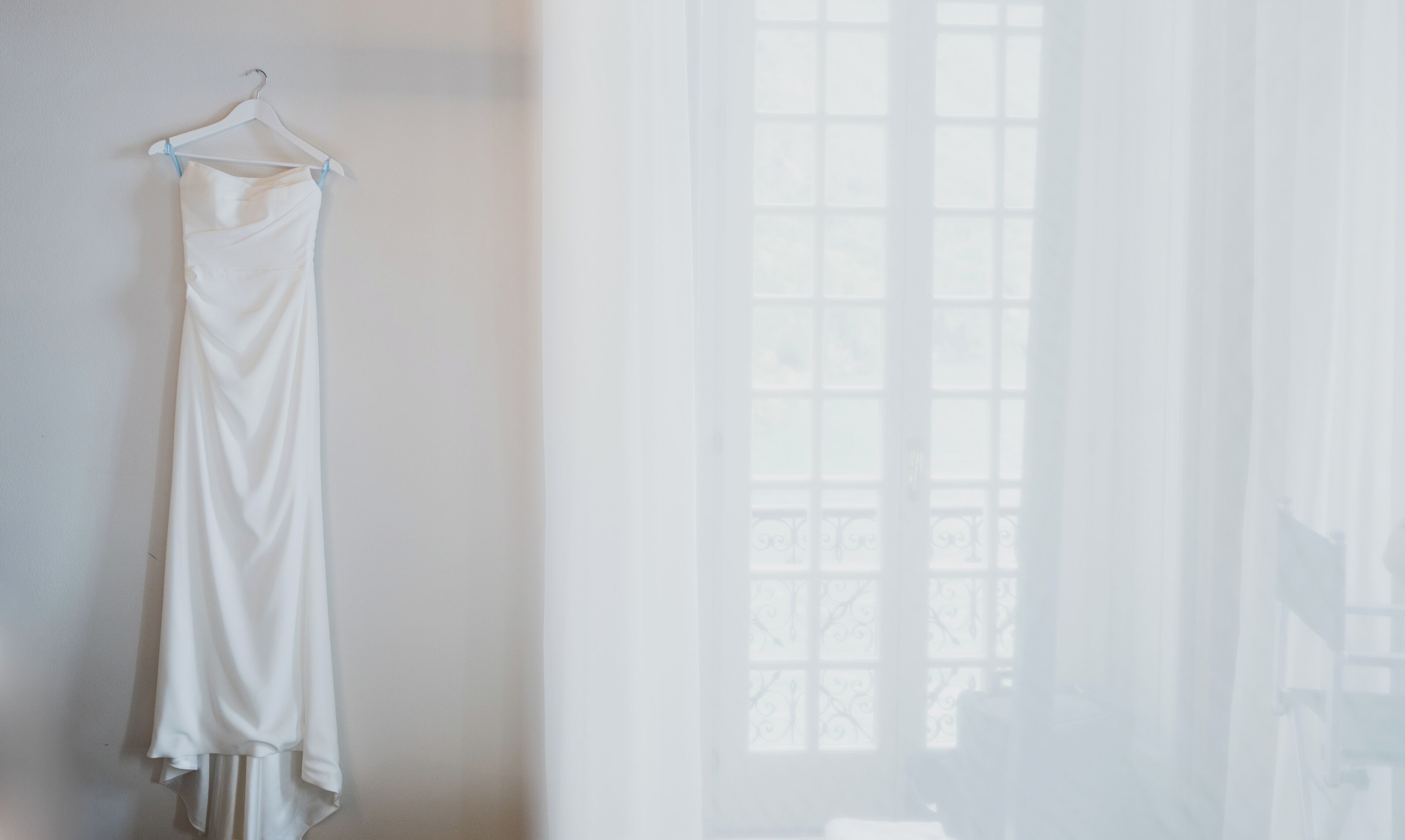 A white dress hanging on a hanger against a wall, with a window in the background.