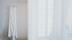 Un vestido blanco colgado en una percha frente a una pared, con una ventana al fondo.