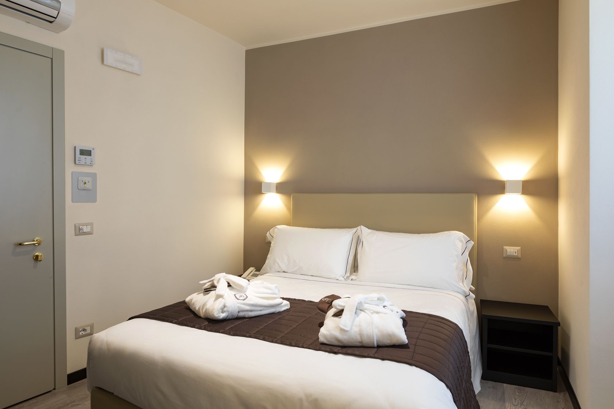 A hotel room with a bed, two wall-mounted lights, a thermostat, and a door with a handle.