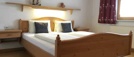 Una cama de madera con cabecera, dos mesitas de noche, una estantería con objetos decorativos, una ventana con cortinas y un cuadro colgado en la pared.