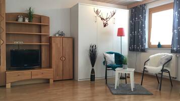 A living room with a wooden cabinet, a television, a small table, two chairs, a lamp, and a window with curtains.