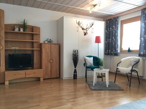 Ein Wohnzimmer mit Holzschrank, Fernseher, einem kleinen Tisch, zwei Stühlen, einer Lampe und einem Fenster mit Vorhängen.