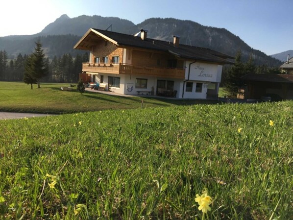 Une maison à deux étages avec un balcon, entourée d’un champ verdoyant et avec des montagnes en arrière-plan.