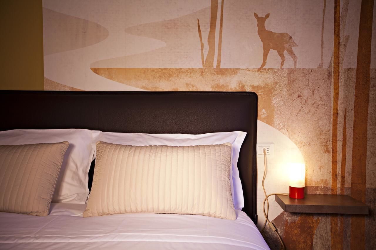 En seng med hvide sengetæpper og to beige puder, et natbord med en rød lampe, samt en væg med silhuetter af en kænguru og træer.