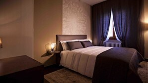 A neatly arranged bedroom with a large bed, a bedside table with a lamp, and a window with curtains.