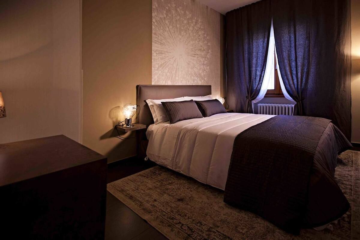 A neatly arranged bedroom with a large bed, a bedside table with a lamp, and a window with curtains.