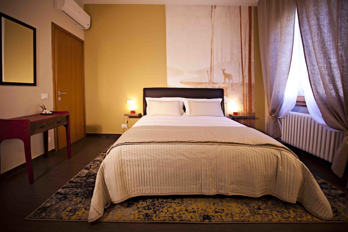 A hotel room with a bed, bedside lamps, a nightstand, a desk, and a large window with curtains.