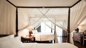 A four-poster canopy bed with a nightstand and lamp, a bedside table with a book and a vase, and a dresser with drawers. - SAFARI PARK HOTEL (NAIROBI)