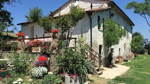A rustic white building with a tiled roof, surrounded by a well-maintained garden with various flowering plants and a wooden fence. - LeAmorine White House B&B (Predappio)