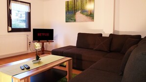 A living room with a brown sofa, a wooden coffee table, a TV displaying 'NETFLIX', a painting of a forest path, and a window with curtains. - Gräfrath Guesthouse Neunkirchen-Seelscheid (Neunkirchen-Seelscheid)