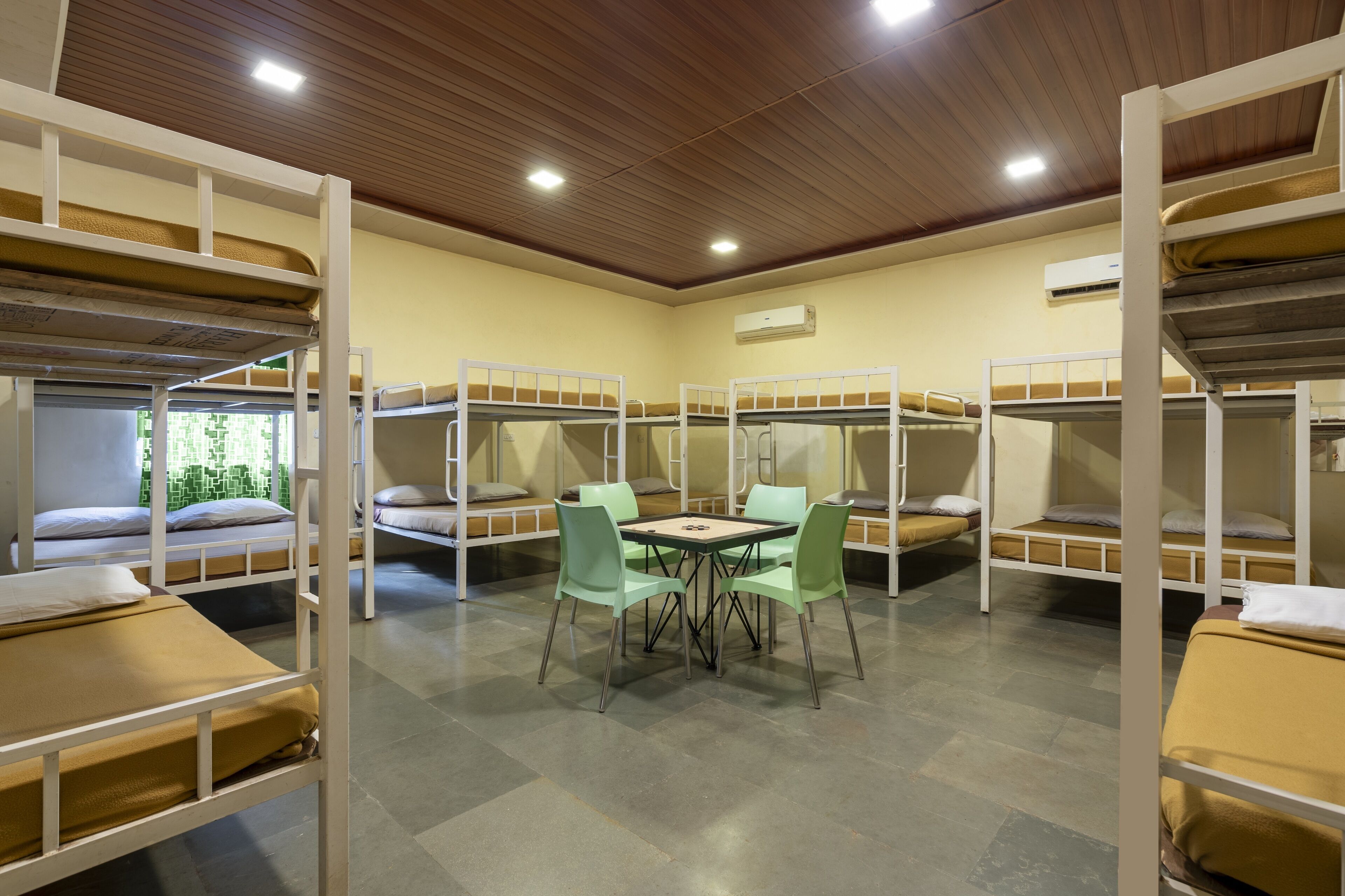 A dormitory room with bunk beds, a table with chairs, and an air conditioning unit on the wall.
