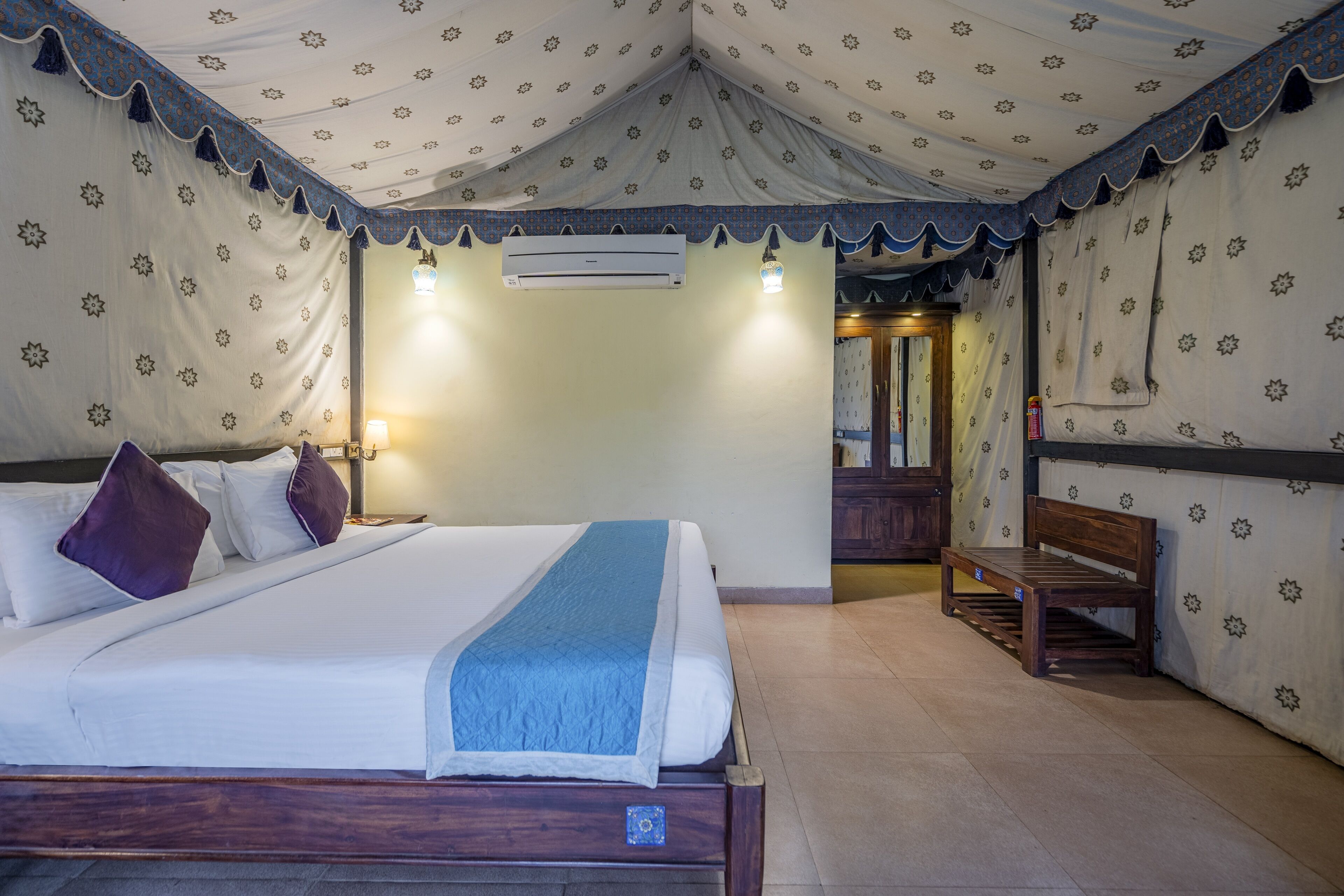 A bedroom with a bed, pillows, a bench, an air conditioning unit, and patterned curtains.