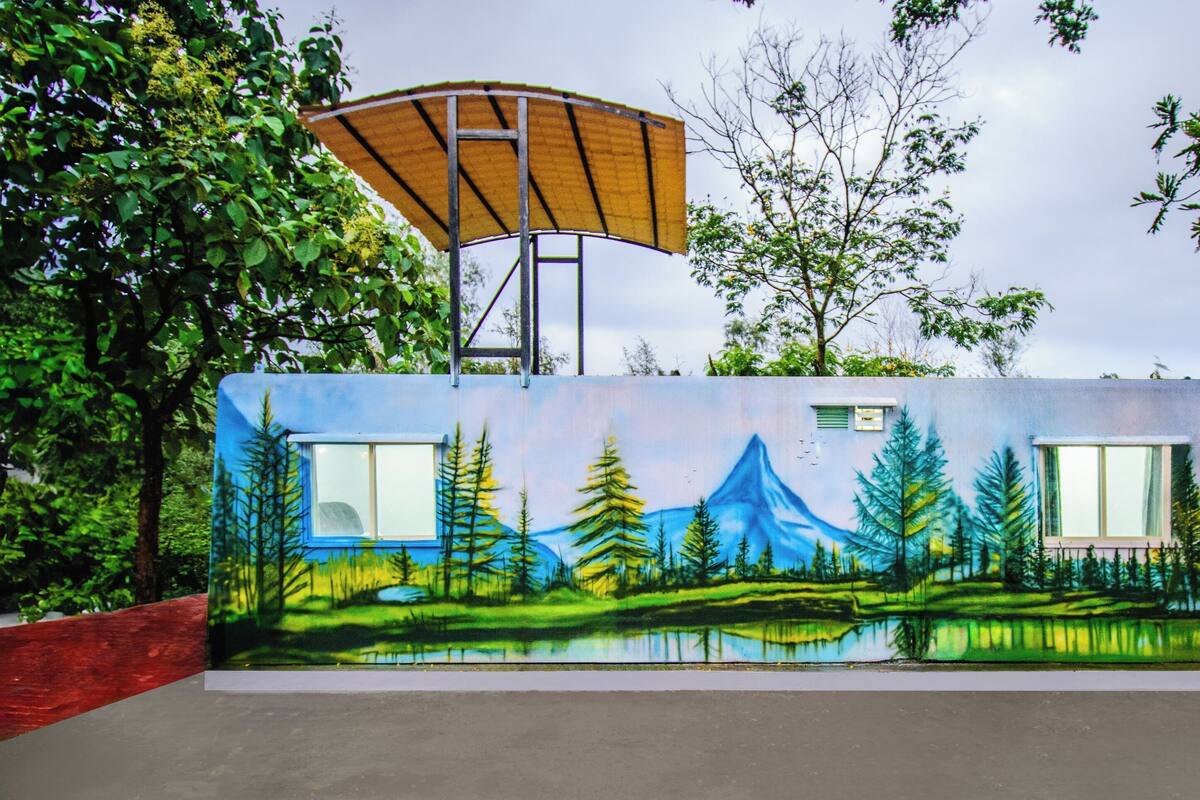 Un edificio con un mural que muestra una escena de bosque, una zona cubierta y dos ventanas.