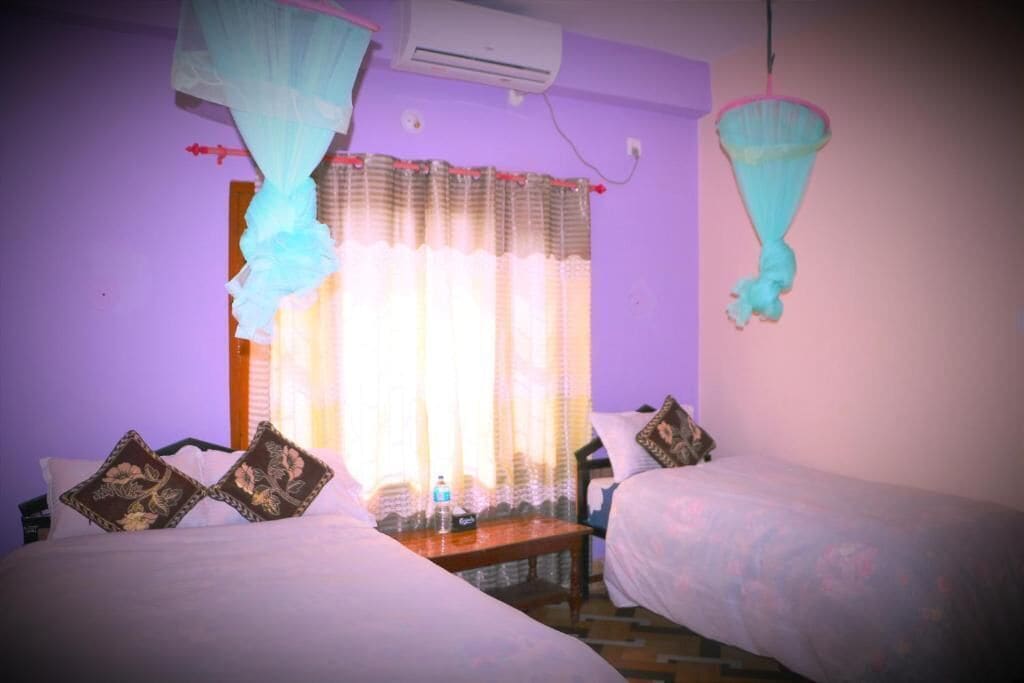 A room with two beds, a bedside table with a water bottle, a mosquito net, and a window with curtains.