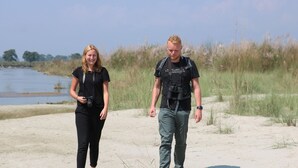 Dos personas caminando por un sendero arenoso cerca de un cuerpo de agua, con césped y árboles al fondo.