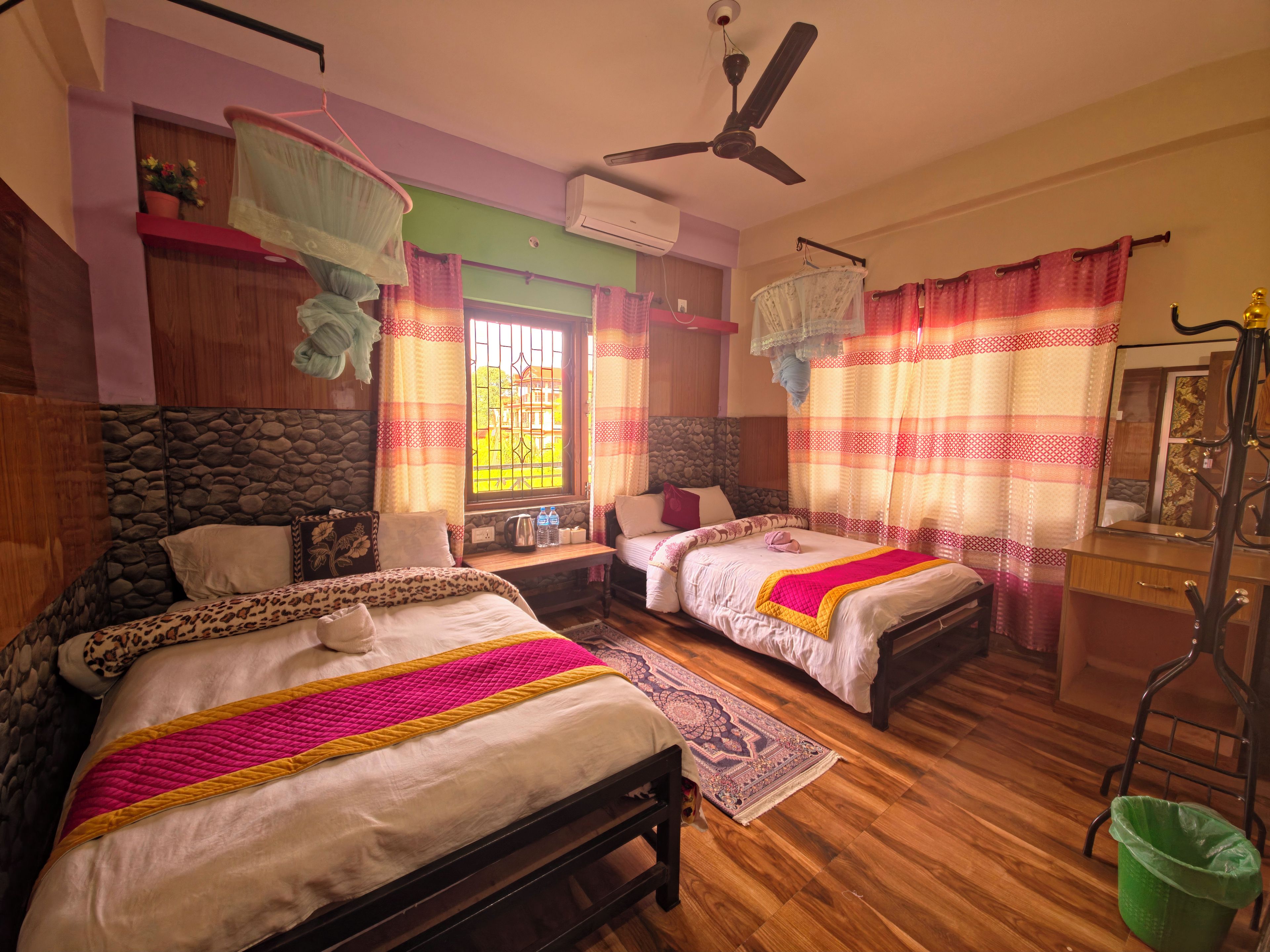 A room with two beds, a ceiling fan, a window with curtains, a green trash bin, and a wooden dresser.