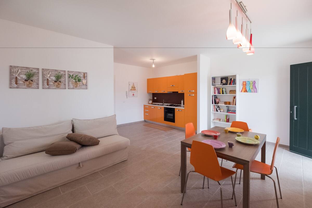 A modern living room with a sofa, dining table, and chairs. There is a kitchen area with orange cabinets and a dining table set with plates and cups.