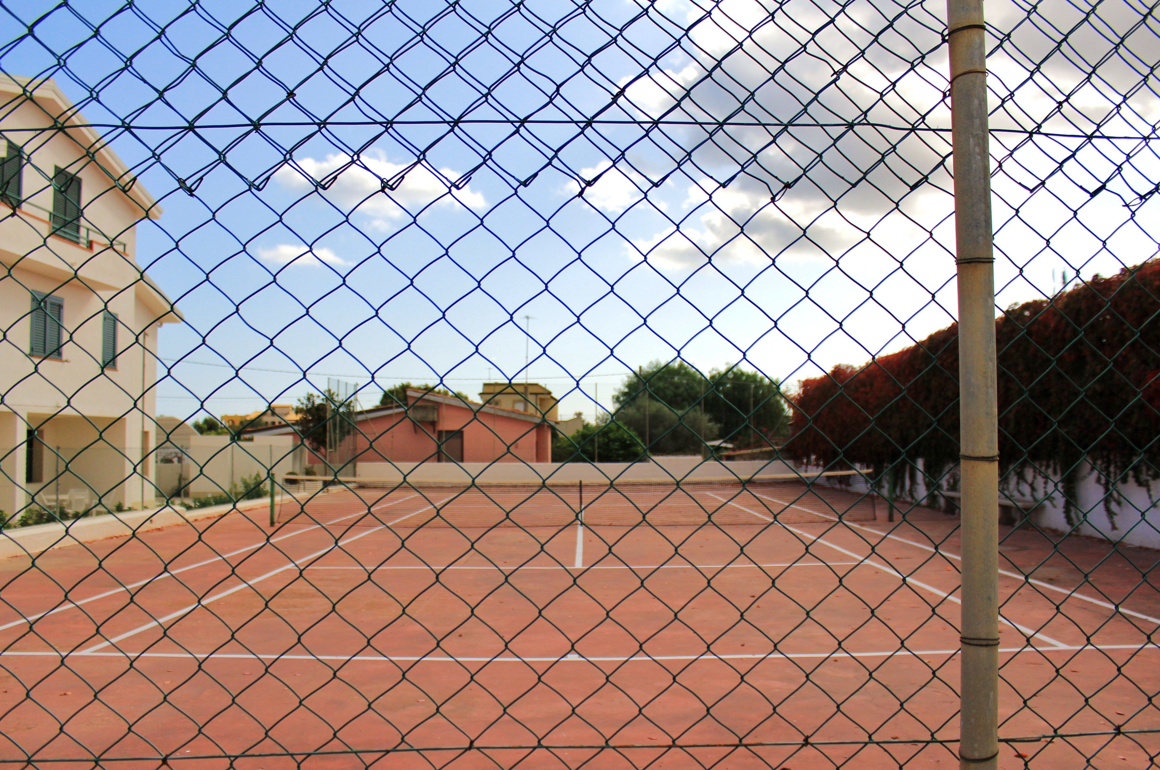 Un court de tennis entouré d’une clôture en treillis métallique, avec un bâtiment en arrière-plan.