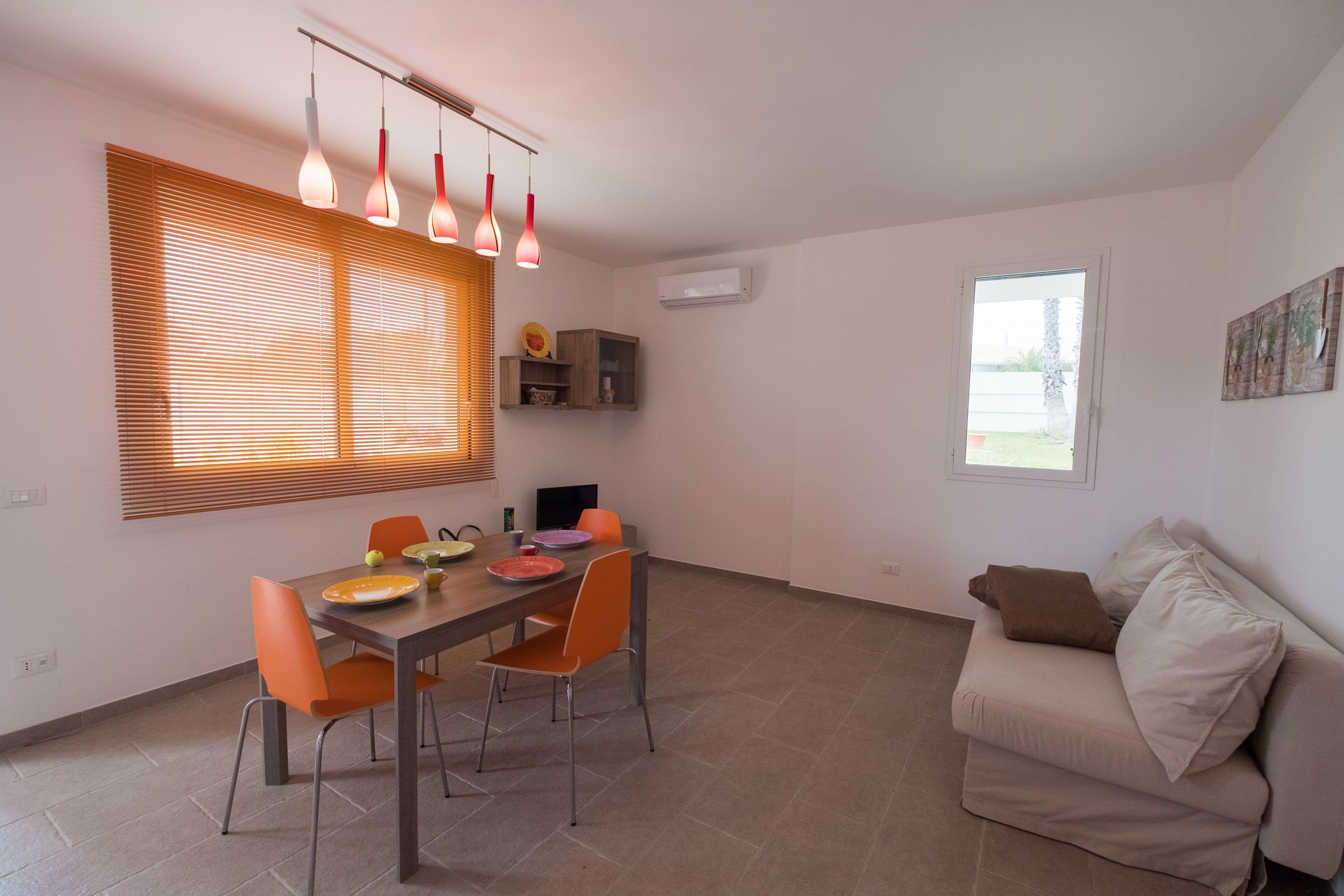 Ein Essbereich mit Tisch, Stühlen und einer Couch. Ein Fenster mit Jalousien, eine Deckenleuchte und ein an der Wand befestigtes Regal mit Dekorationen sind vorhanden.