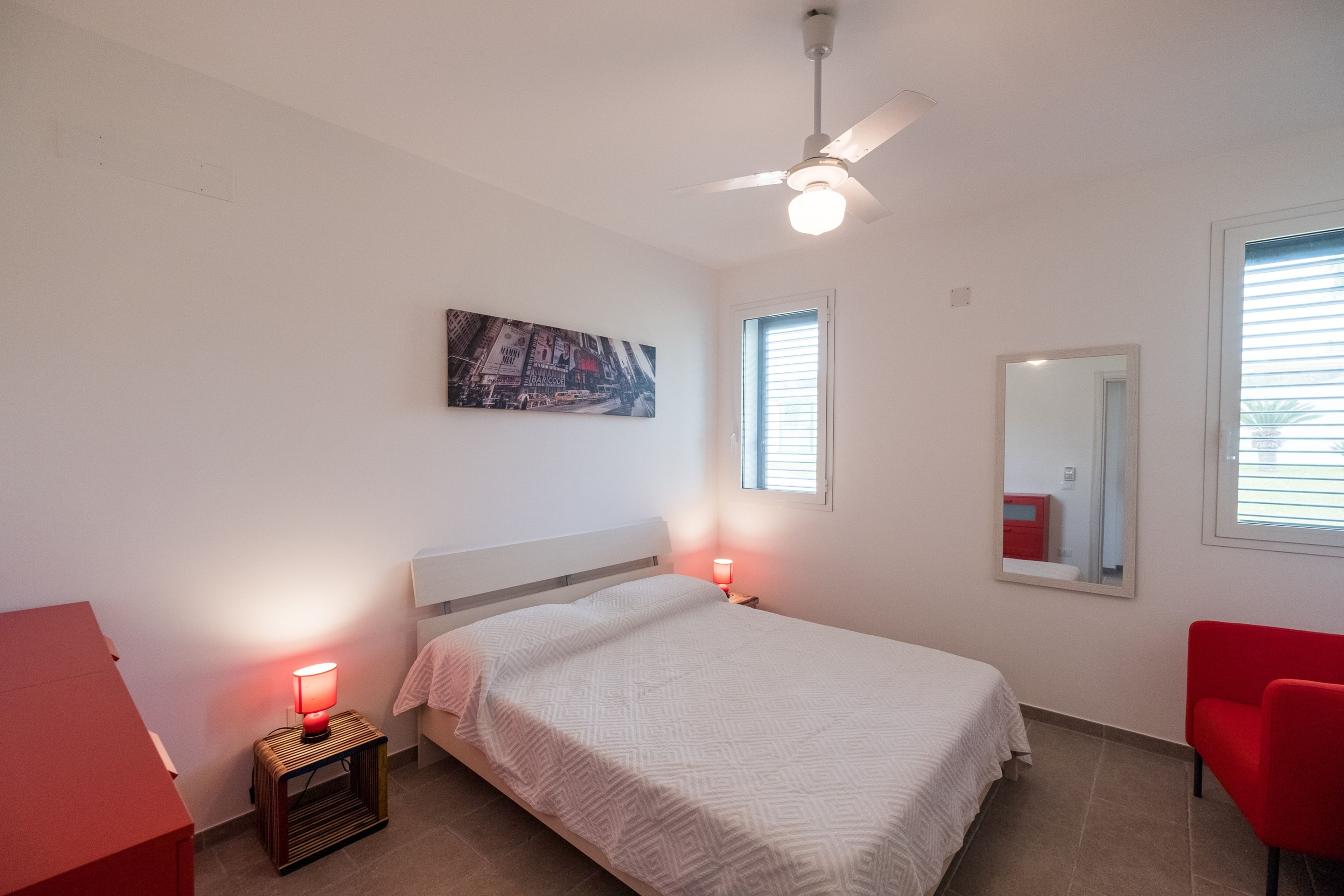 A bedroom with a bed, a red chair, a nightstand with a lamp, a ceiling fan, a window with blinds, and a painting on the wall.