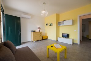 Ein modernes Wohnzimmer mit einer Couch, einem gelben Tisch mit einem Buch und einem Telefon, einem Fernseher auf einer weißen Kommode, einer grünen Tür und einer gelben Wand mit Dekorationen.