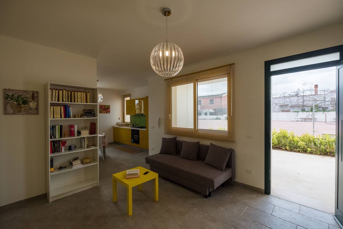 A living room with a sofa, a yellow coffee table, a bookshelf, and a large window with blinds.