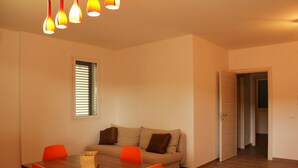Een eetgedeelte met een houten tafel, oranje stoelen, een beige bank en een raam met zonwering.