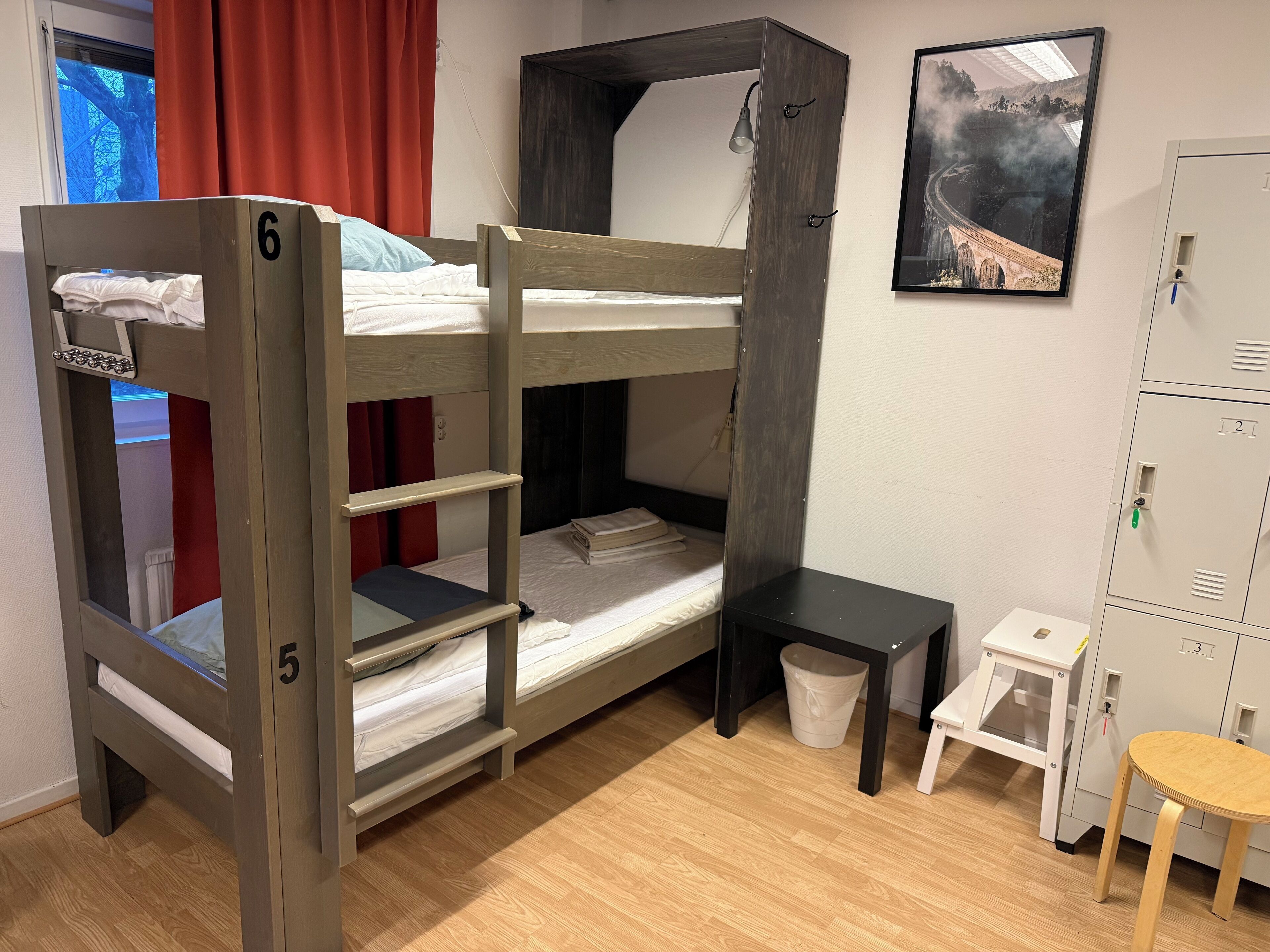 A bunk bed with a desk and a small stool, a waste bin, and a framed picture on the wall.