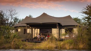 A safari lodge with a covered outdoor seating area overlooking a river.