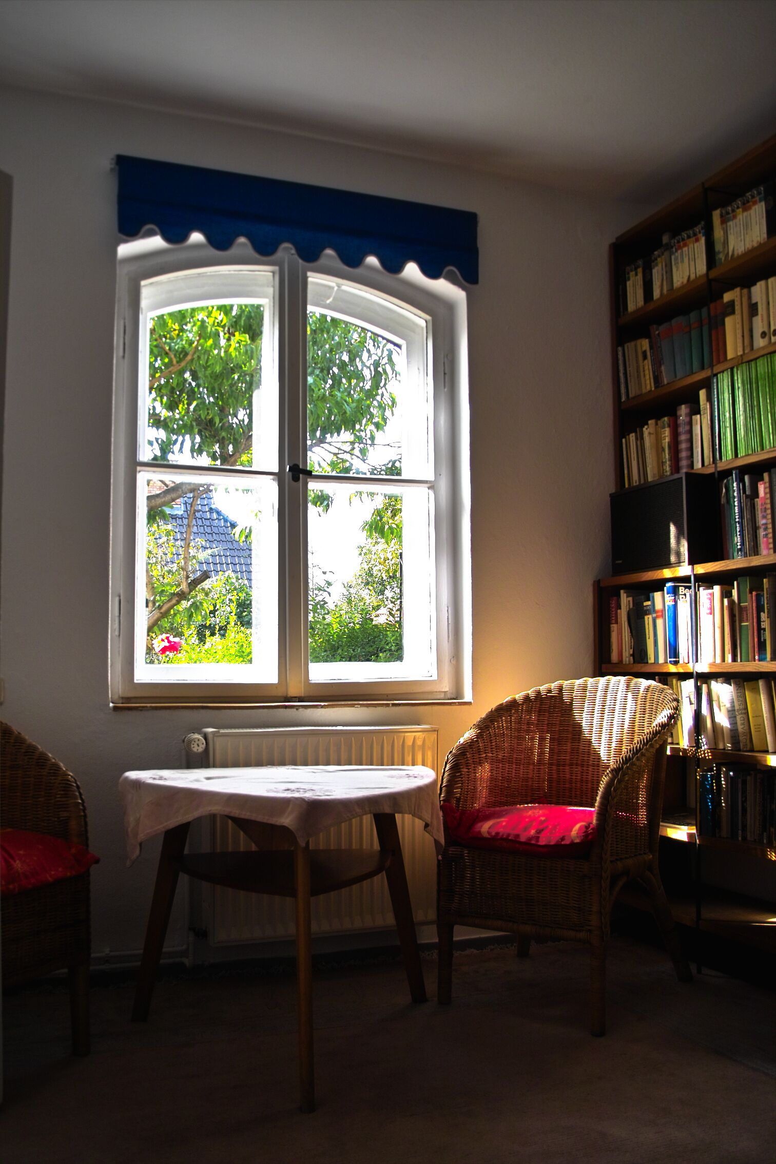 Een kamer met een groot raam, een boekenkast, een rieten stoel en een tafel.