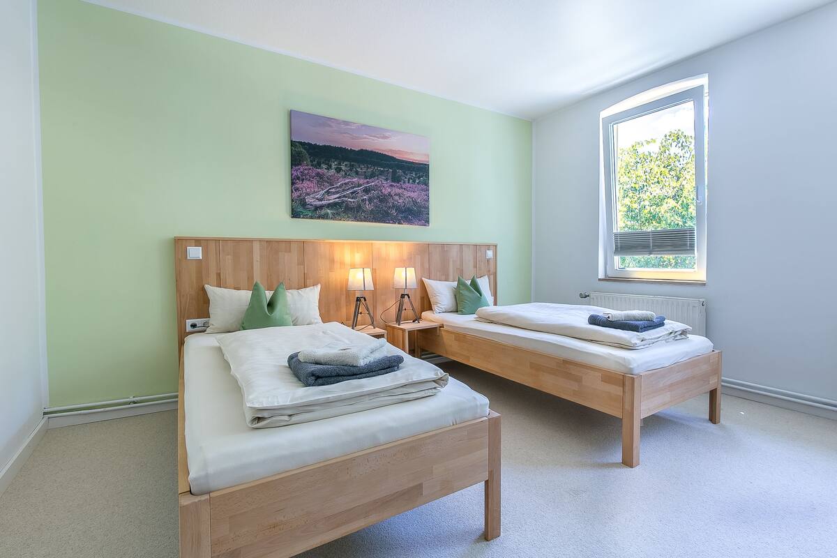 A bedroom with two wooden beds, a nightstand with lamps, a painting on the wall, and a window with a view of trees.