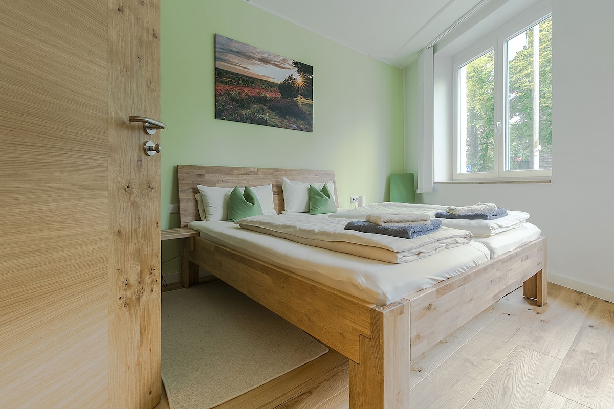 A bedroom with a wooden bed, a green wall, and a window with a view of trees.