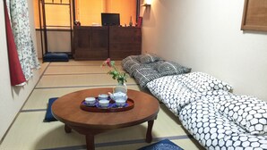 Un salón japonés tradicional con suelo de tatami, una mesa de madera baja con un juego de té, un sofá con cojines estampados y un televisor sobre un pedestal.