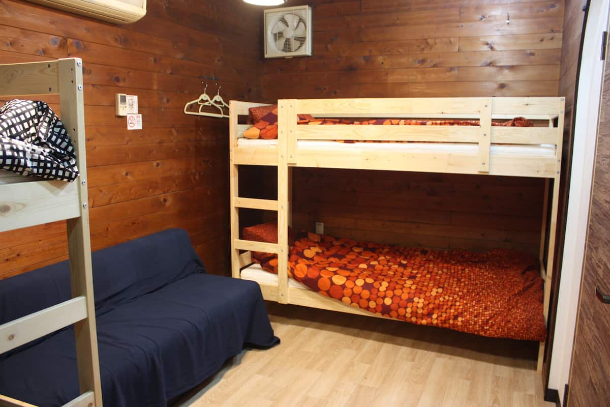 Una cama litera con la parte inferior llena de almohadas naranjas, un sofá azul y una pared de madera con un ventilador y un interruptor de luz.