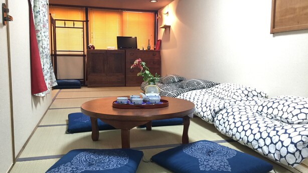 Una habitación con suelo de tatami, una mesa baja con una bandeja de objetos, un sofá con cojines estampados y un armario de madera al fondo.