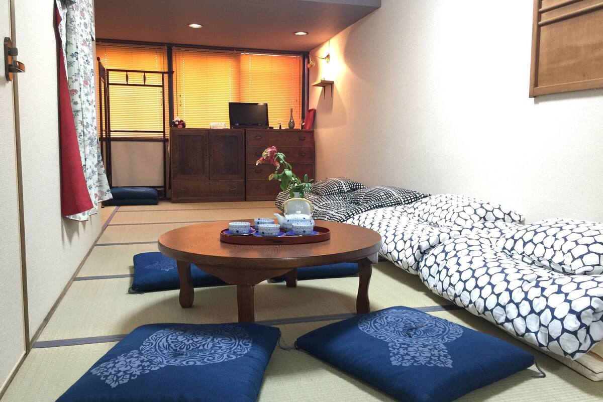 Una habitación con suelo de tatami, una mesa baja con una bandeja de objetos, un sofá con cojines estampados y un armario de madera al fondo.