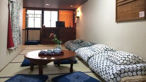 Una habitación tradicional japonesa con suelo de tatami, una mesa baja con juego de té, un sofá con cojines estampados y una puerta corredera que da a un balcón.