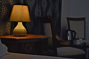A lamp on a wooden nightstand in a dimly lit room with patterned curtains and a chair. - PJ Lodge (Dar es Salaam)